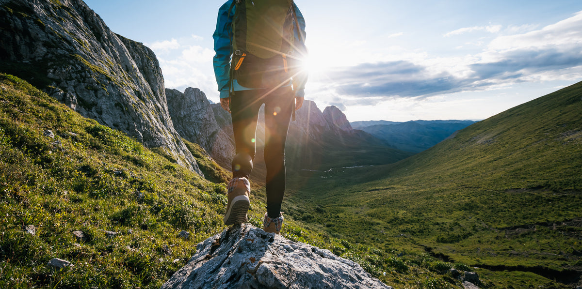 A hiker wearing a blue jacket and black backpack stands on a rocky peak in a mountain valley, with the sun shining brightly behind them and green hills stretching into the distance.